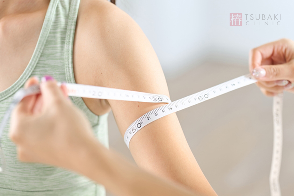 ノースリーブのトップスを着た女性の二の腕を、スタッフがメジャーで丁寧に採寸している様子。腕まわりのサイズを確認するため、メジャーが二の腕に沿って巻かれている画像