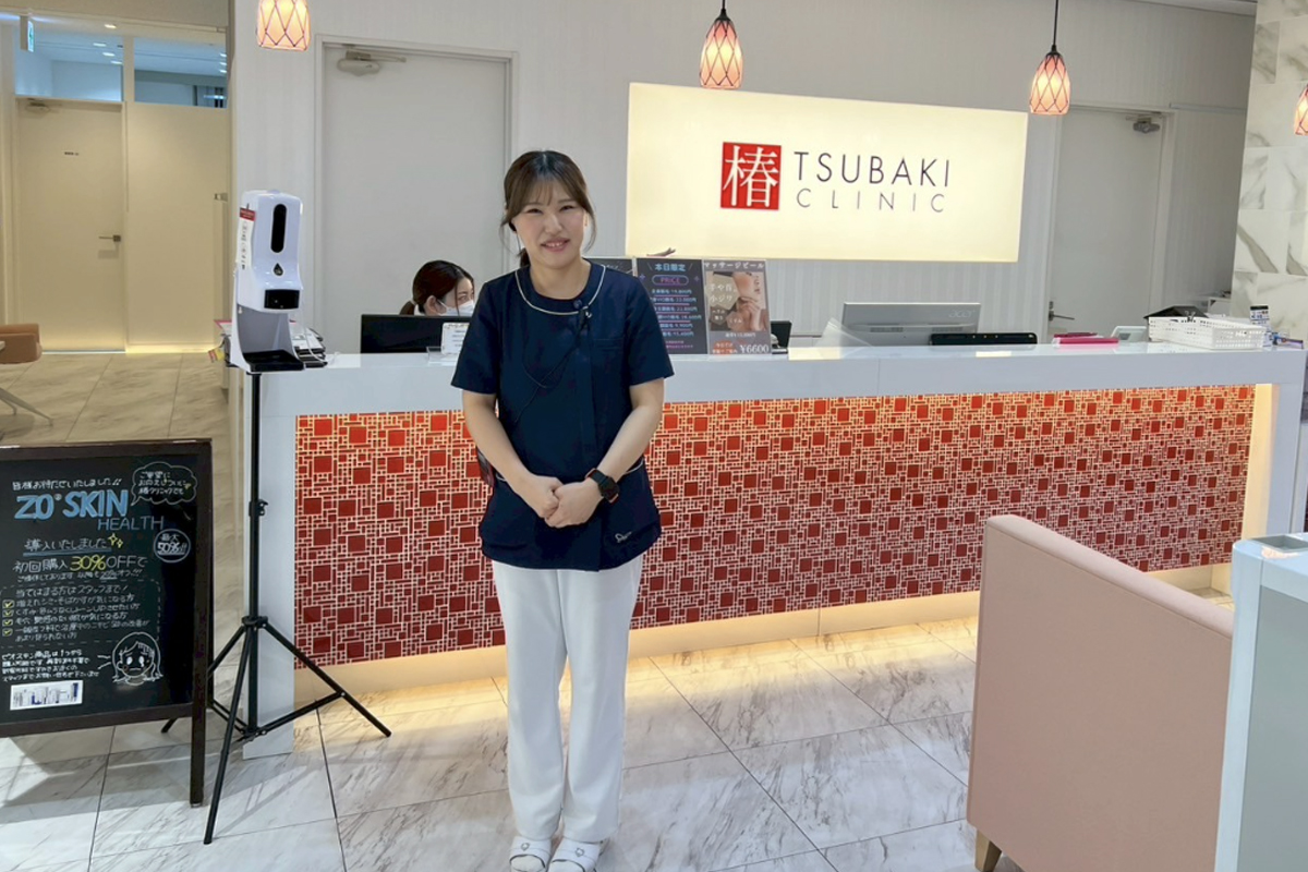 椿クリニックの入口で女性スタッフがお客様を案内する風景の実写写真。赤と白を基調とした受付で、その背景には椿クリニックの表記がある。