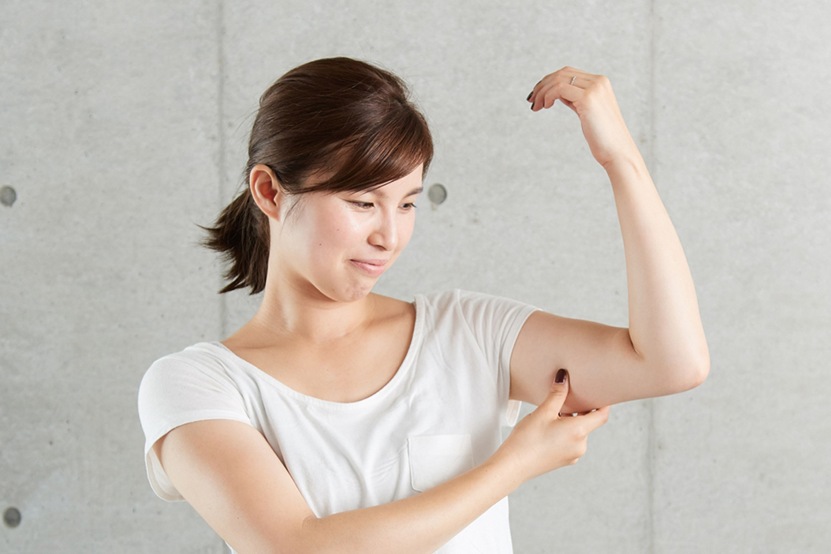 白いTシャツを着た女性が腕を曲げ、二の腕の内側を指でつまんでたるみを確認している様子。気になる部分をチェックしながら少し困った表情を浮かべている写真。