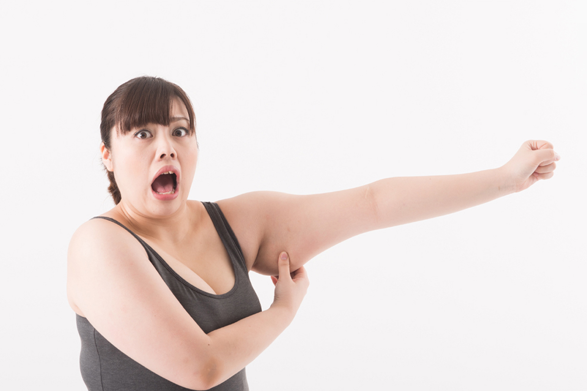 少しふくよかな体系のタンクトップ姿の女性が腕を横に伸ばし、二の腕のたるみをつまんで驚いた表情をしている様子。自分の二の腕のたるみを予想以上に感じて戸惑っているイメージ写真。