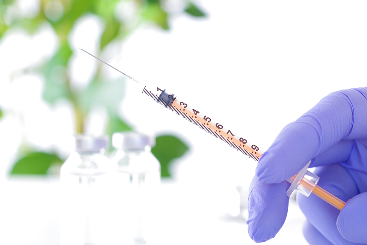 注射器を持った手袋をした手元のアップ。背後には薬液の入ったバイアルが複数置かれている。