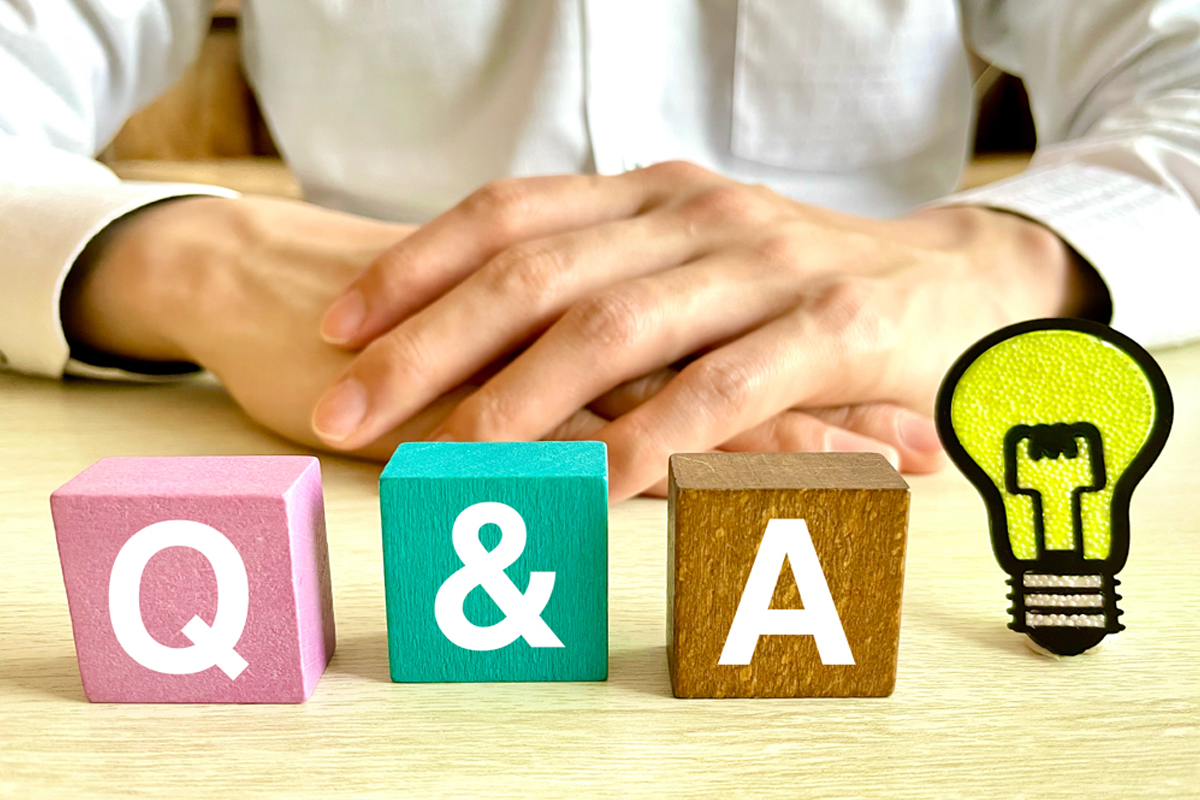「Q&A」と書かれたブロックと電球のイラストがテーブルに置かれている。背後にはシャツを着た人物が座っており、重ねた手の部分だけ写っている。