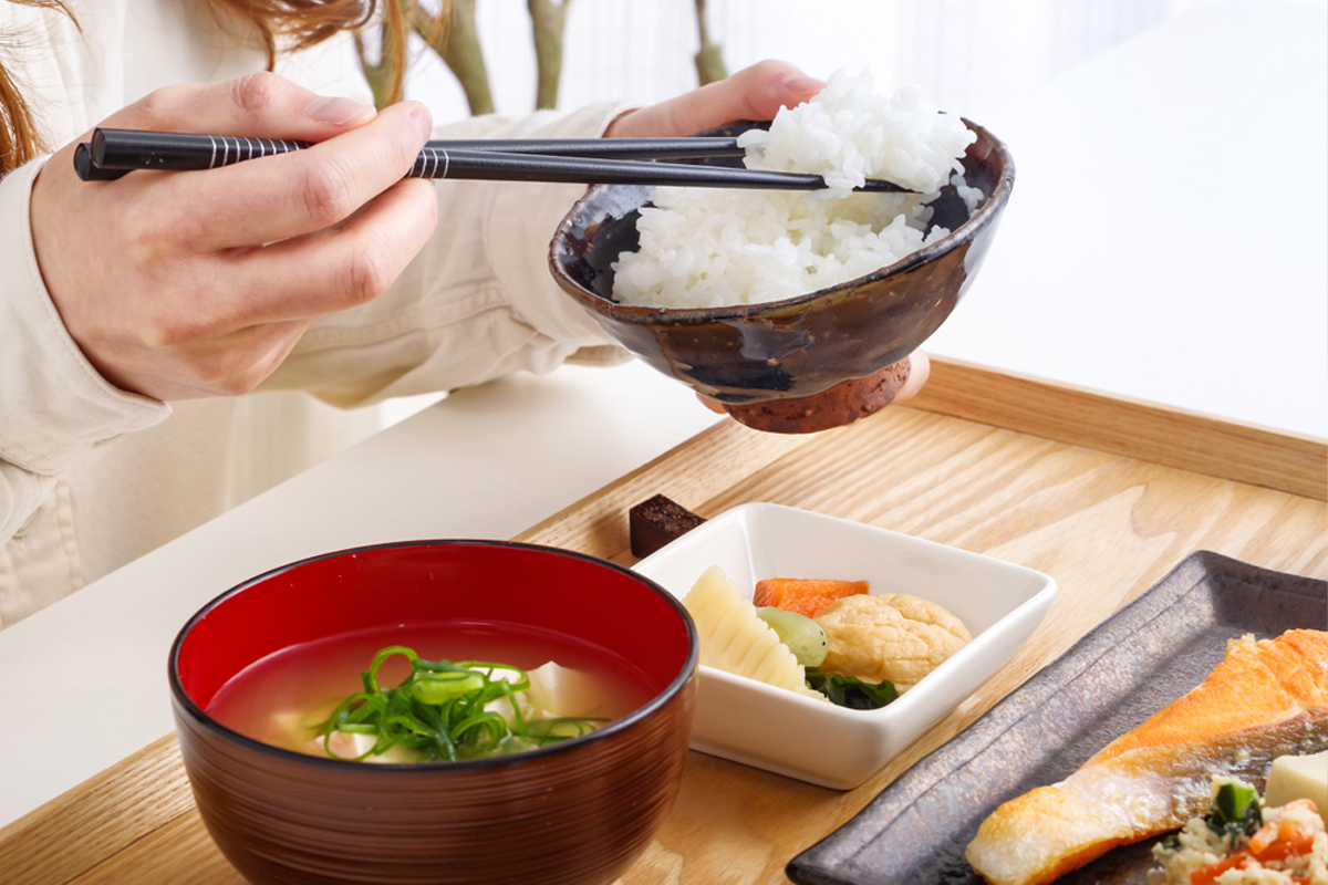 茶碗に入ったごはんを箸で持ち上げ、手前にはみそ汁、奥には焼き鮭と小鉢が並んでいます。