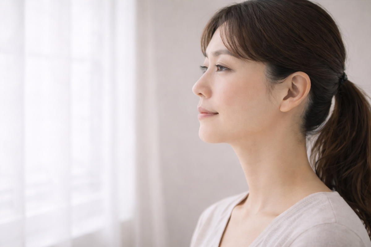 フェイスラインの美しい40代女性の横顔。明るい窓際にたたずんでいる。その表情は穏やか。