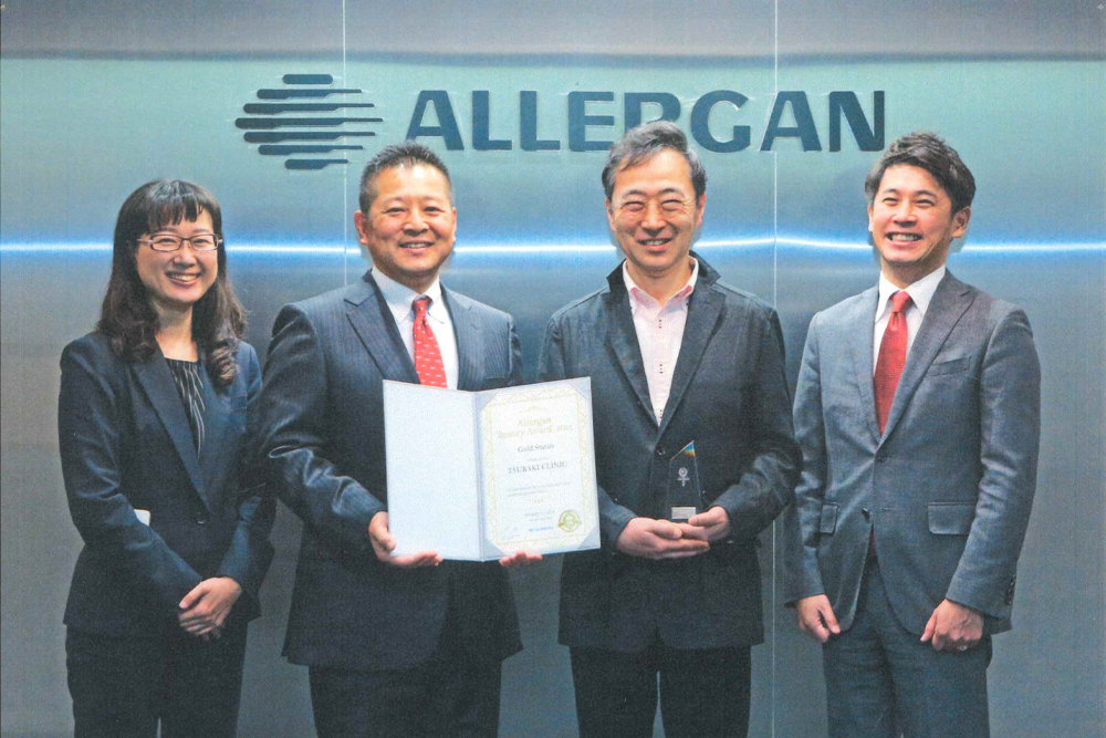 「ALLERGAN」と書かれた壁の前で、スーツ姿の3名と椿クリニックの田辺理事長が並び、認定証とトロフィーを手にしている集合写真。